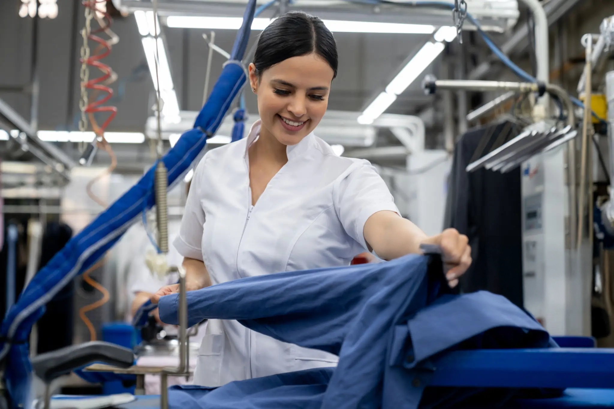 A woman in a white uniform irons a blue shirt in a commercial laundry or dry cleaning facility.