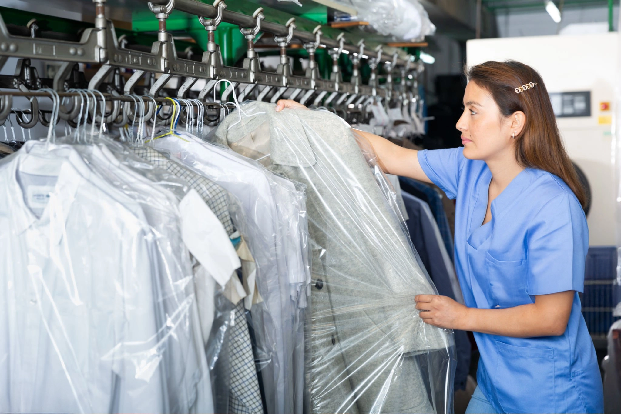 Woman in blue uniform selecting garments covered in plastic from a rack at a dry cleaning facility.