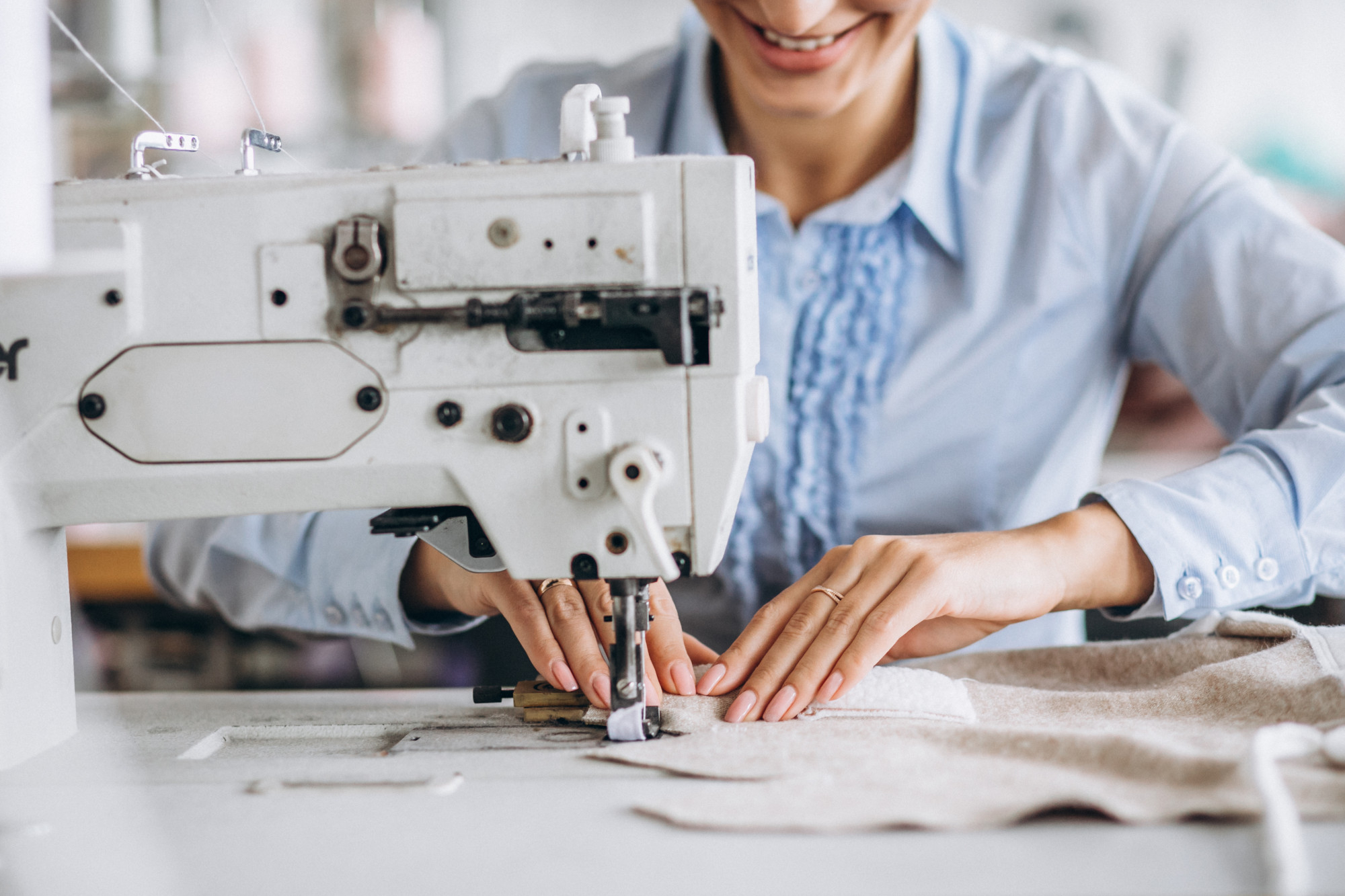 A person uses an industrial sewing machine to stitch light-colored fabric, smiling and concentrating on their work.