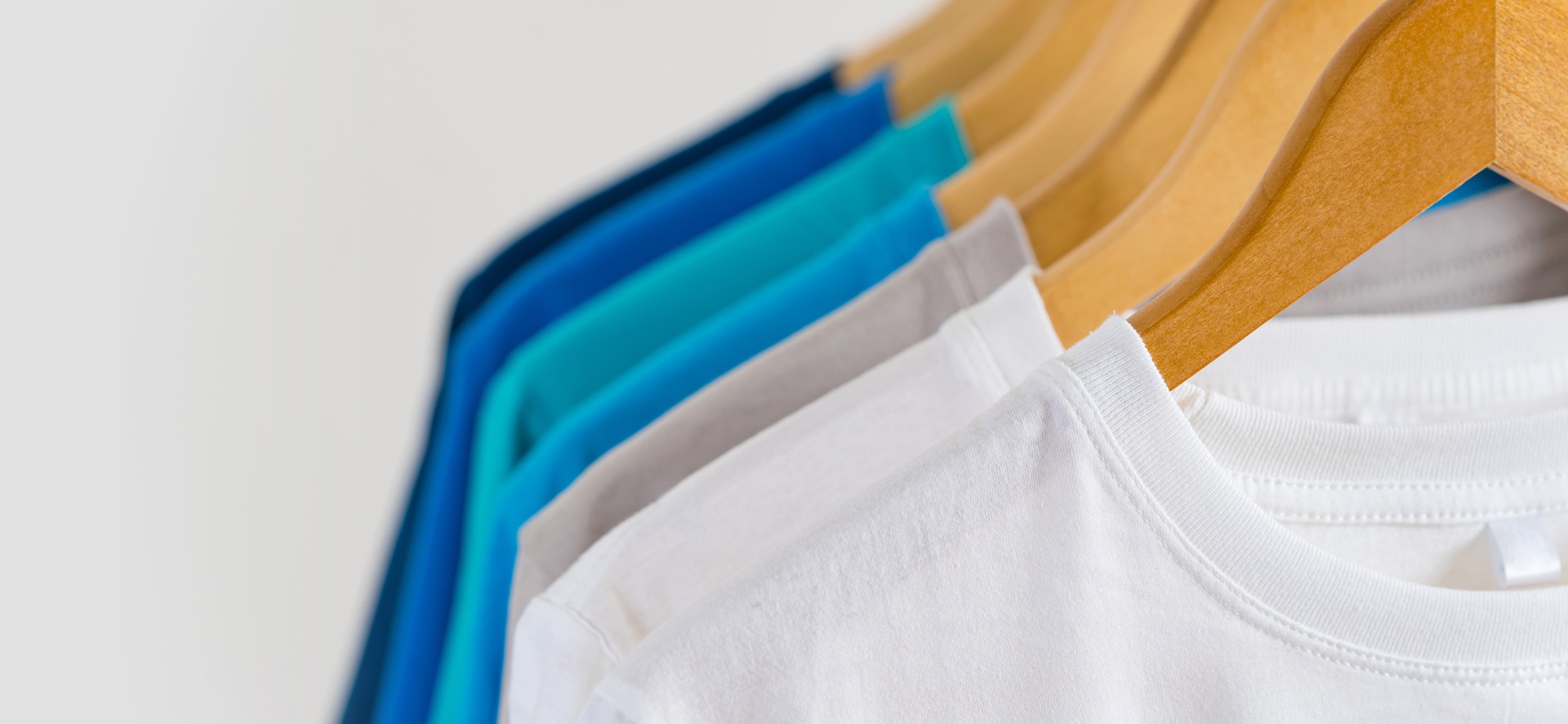 A row of plain t-shirts in white, gray, and shades of blue hangs neatly on wooden hangers against a light background.