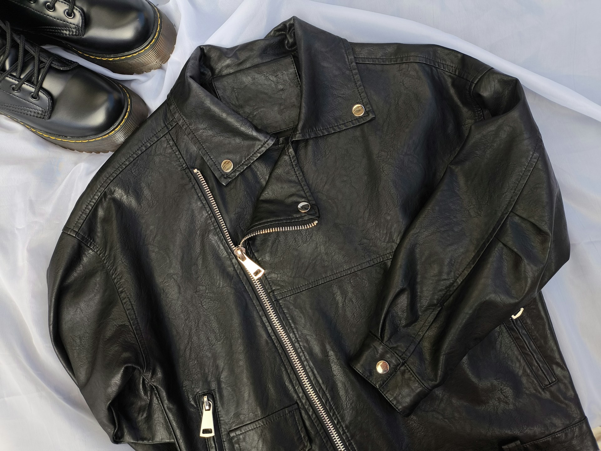 Black faux leather biker jacket with silver snaps and zippers laid out on a light fabric, next to a pair of black lace-up boots with thick soles.