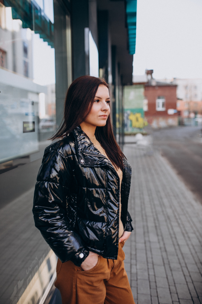 A woman with long dark hair stands on a city sidewalk, wearing a black shiny jacket and brown pants, with her hands in her pockets.