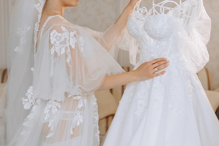 A bride in a sheer, floral-embroidered robe and veil touches a white wedding dress displayed on a mannequin.