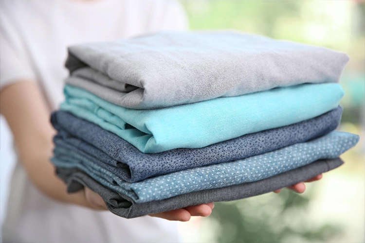 A person holds a neatly folded stack of blue and gray clothes with both hands, against a blurred green background.