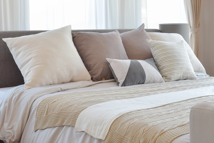 A neatly made bed with white and beige bedding, decorated with several neutral-colored pillows, in a bright room with sheer curtains.