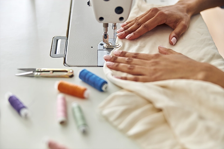 A person uses a sewing machine to stitch light-colored fabric, with spools of thread and scissors on the table nearby.
