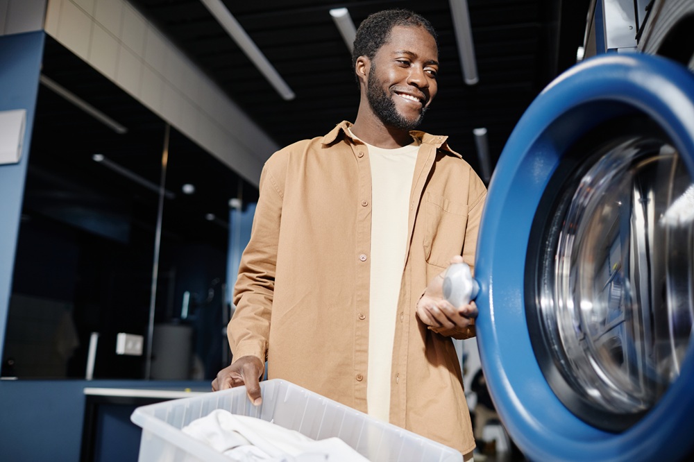 A man holding a laundry basket loads clothes into a front-loading washing machine in a laundry room.