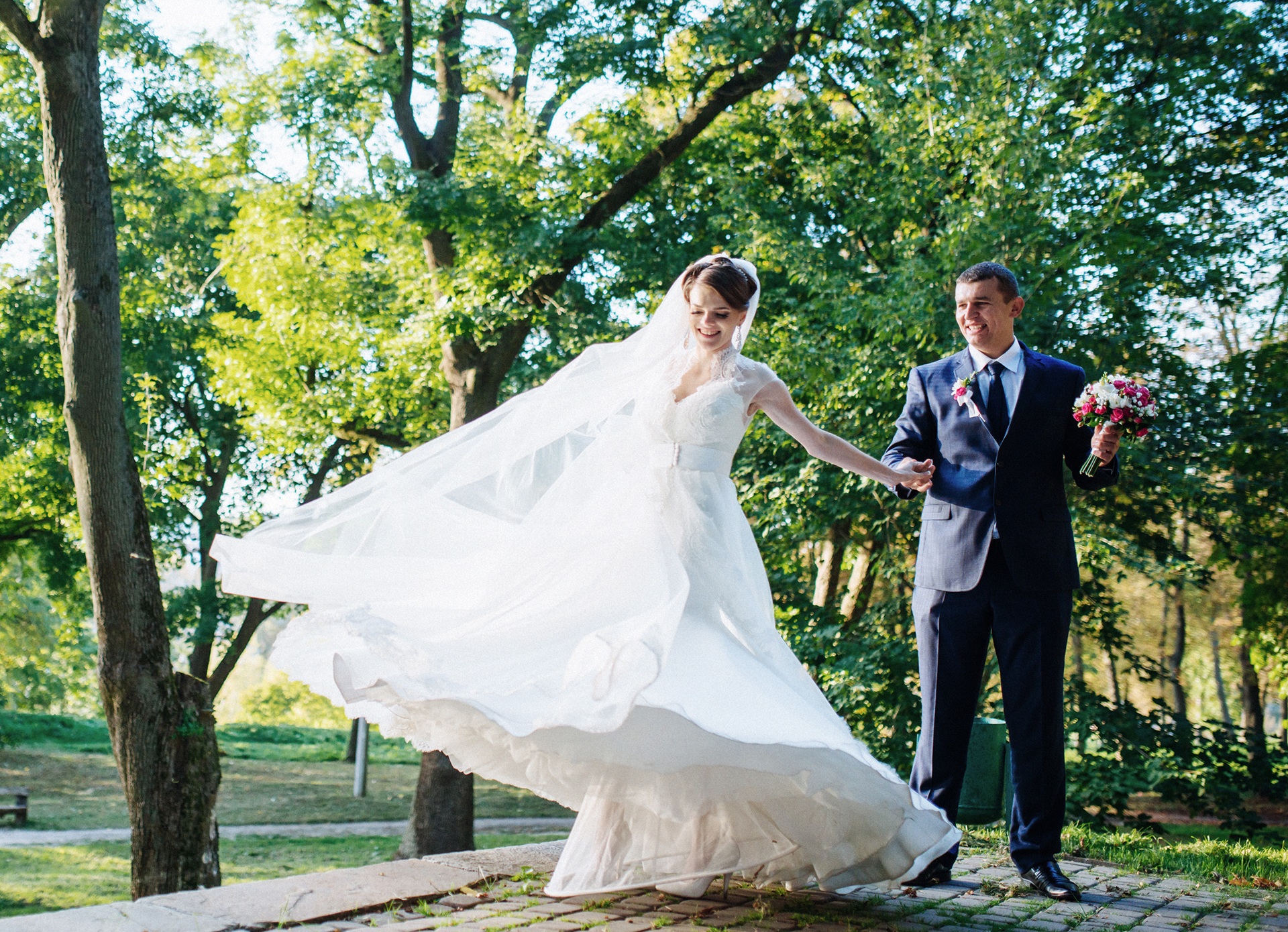 A bride in a white wedding dress twirls next to a groom in a dark suit holding a bouquet, outdoors in a sunlit park with green trees.