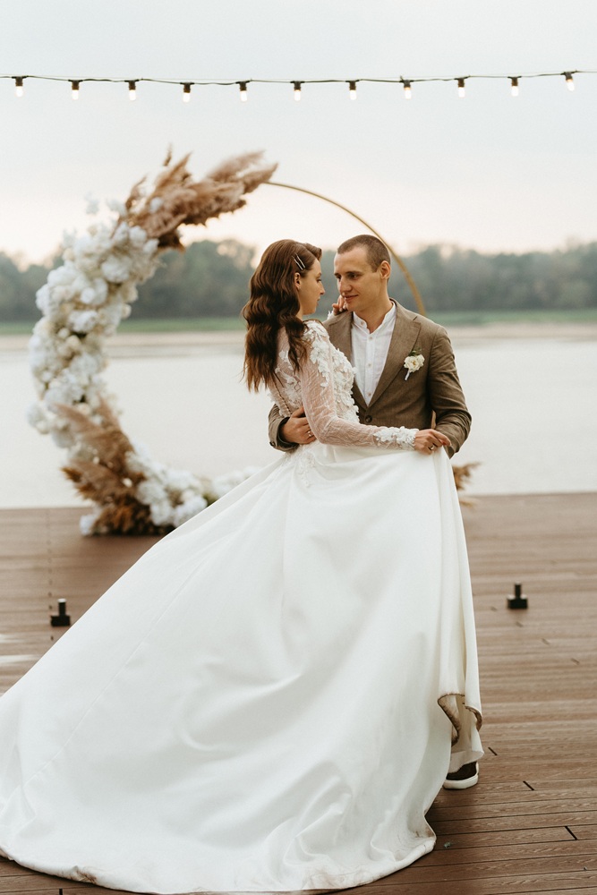 A bride and groom stand on a wooden deck by the water, embracing in front of a circular floral arch.