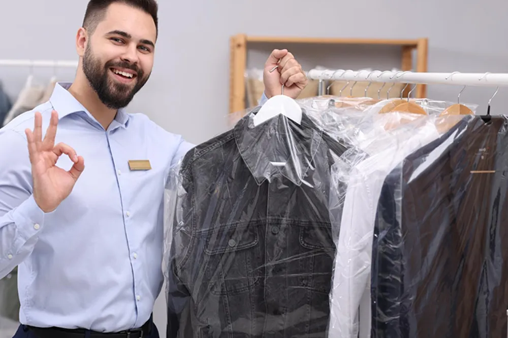 Man in a dress shirt holding cleaned clothes on hangers covered in plastic, standing next to a clothing rack, smiling and making an "OK" gesture.